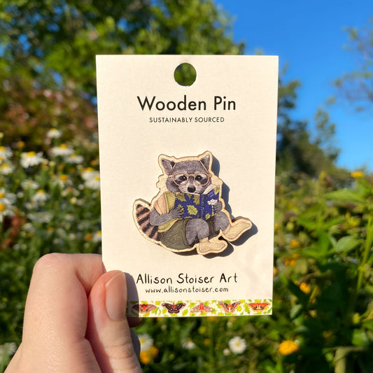 Wooden pin of a raccoon reading a book on a card held in front of a natural background