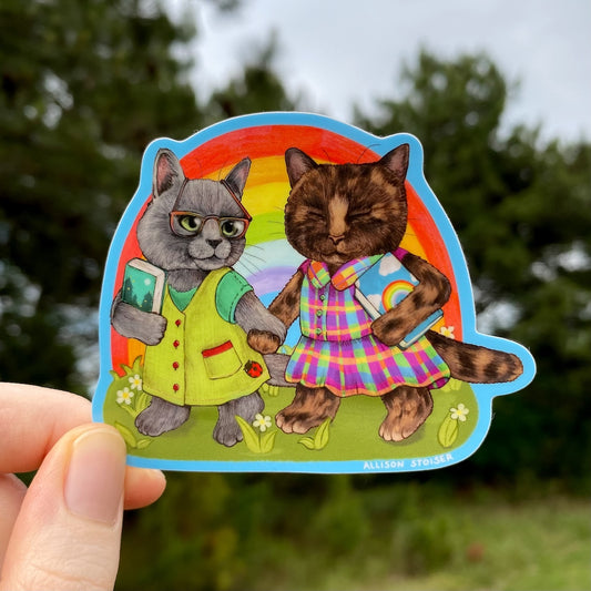Sticker of two cats holding hands with a rainbow background, held by a hand outdoors.