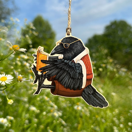 An ornament of a raven wearing glasses and a red-and-white sweater, holding a book. The ornament is photographed outdoors against a blurred green field with white flowers and blue sky.