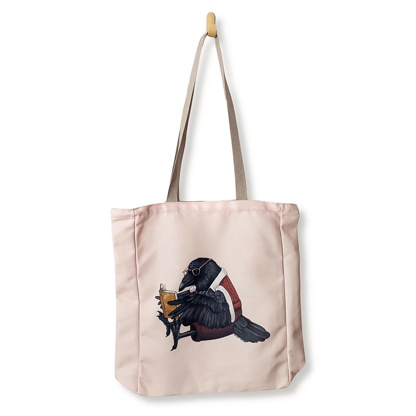 A light pink canvas tote bag with beige shoulder straps, featuring an illustration of a black raven wearing a red sweater vest and holding a yellow book, shown hanging from a wooden hook against a white background.