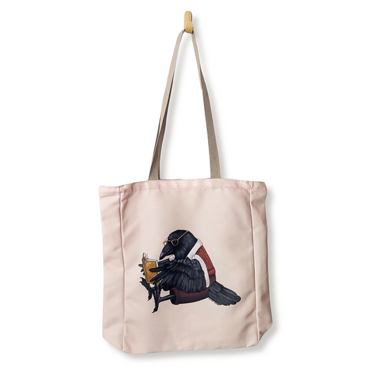 A light pink canvas tote bag with beige shoulder straps, featuring an illustration of a black raven wearing a red sweater vest and holding a yellow book, shown hanging from a wooden hook against a white background.
