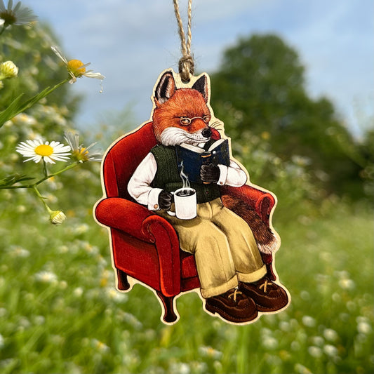 A hanging ornament of a red fox sitting in a red armchair and reading a book, photographed outdoors against a blurred green field with flowers and blue sky.