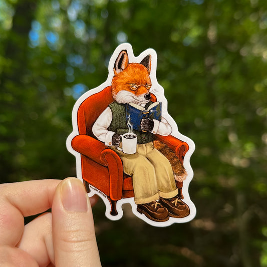 Sticker of a fox sitting in an armchair with a book and coffee, held against a blurred forest background.