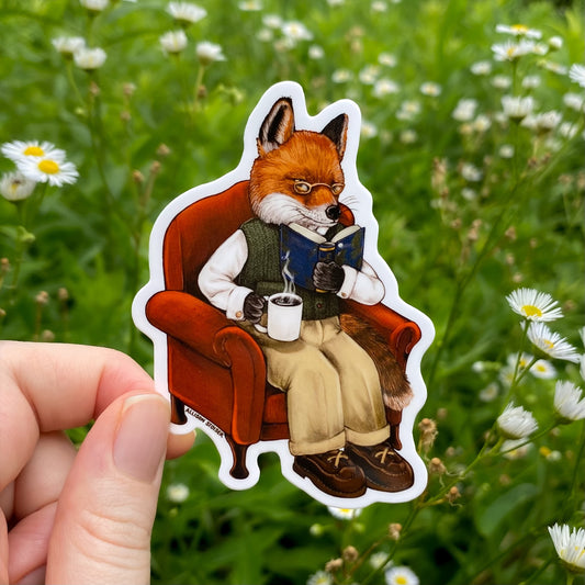 Sticker of a fox reading a book in a chair held in front of a natural background with flowers.