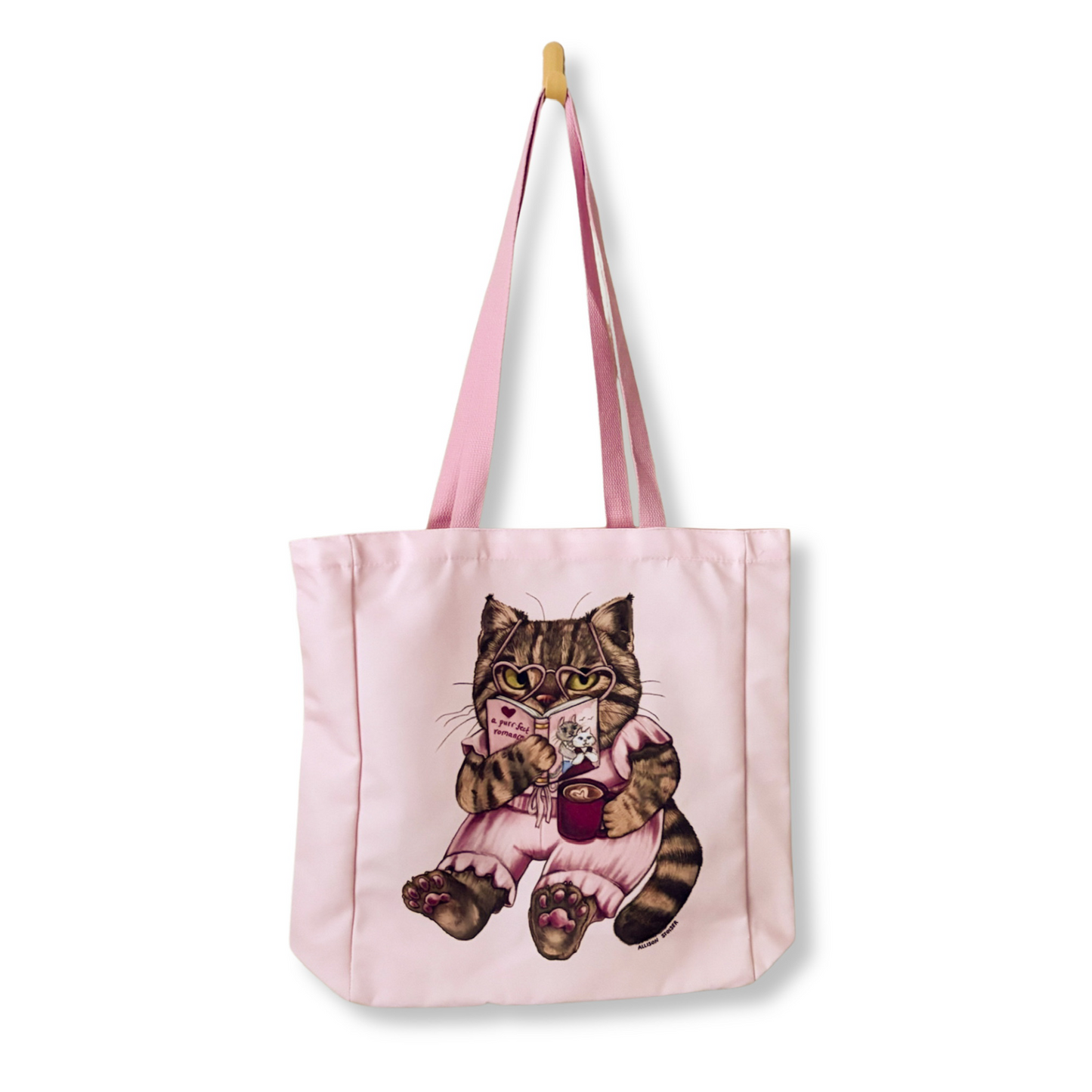 A light pink canvas tote bag with pink shoulder straps, featuring a printed illustration of a tabby cat in a pink outfit holding a romance book and red mug. The bag is shown hanging from a wooden peg against a plain white background.