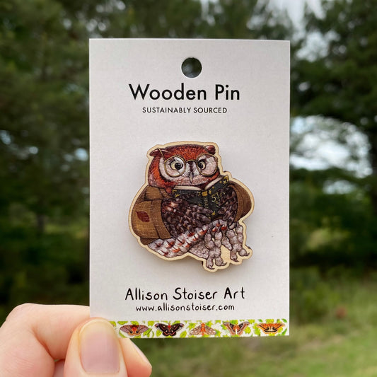 Wooden pin of an owl reading a book on a card labeled 'Allison Stoiser Art' against a blurred natural background.