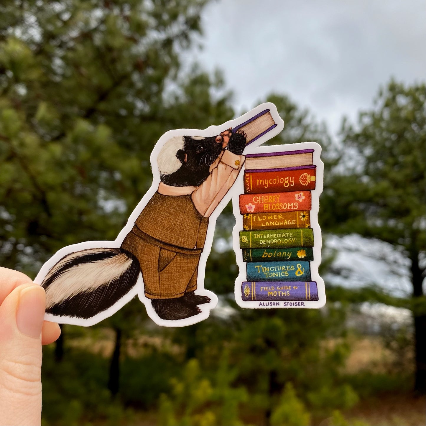 Sticker of a skunk in a tweed vest holding a book with a blurred natural background