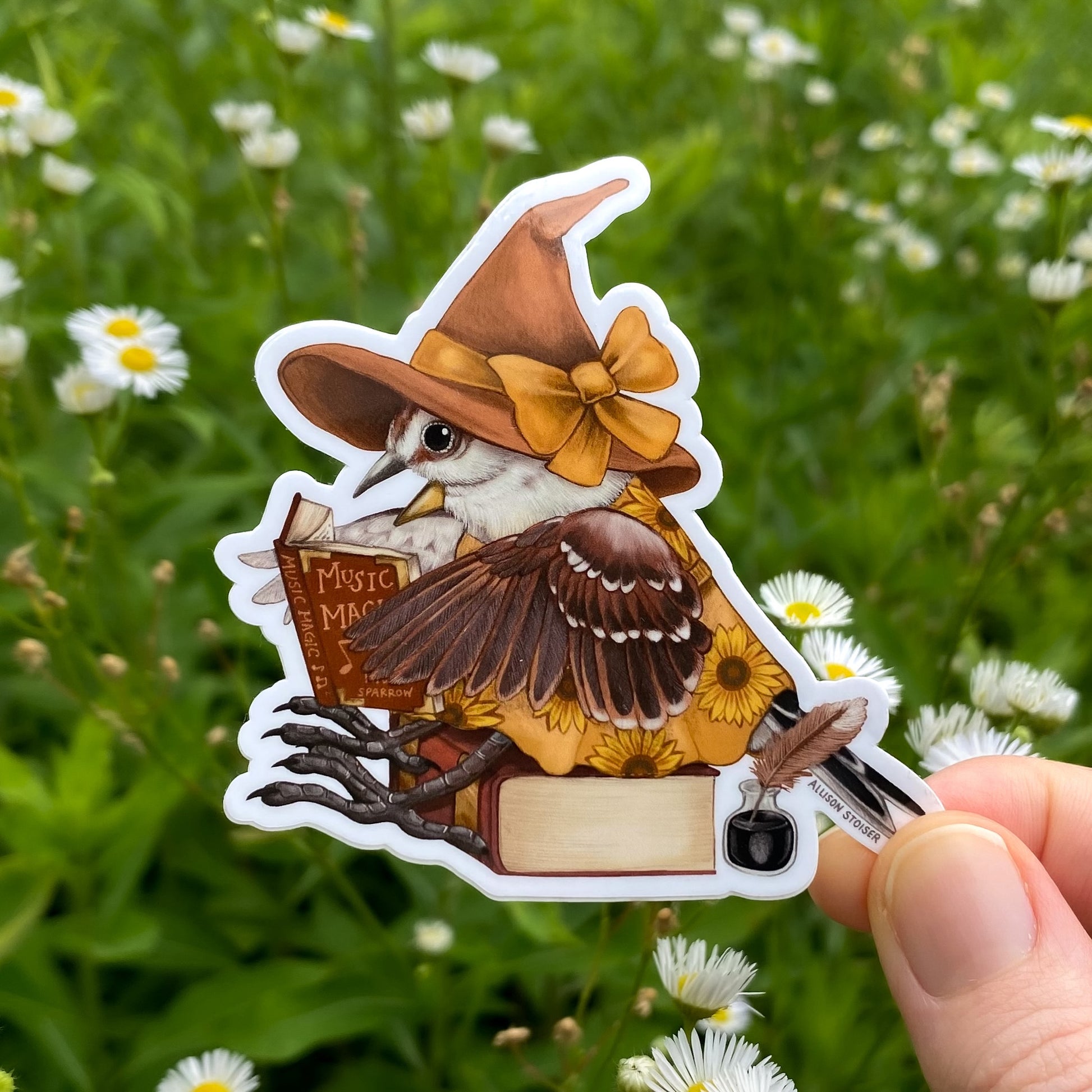 Sticker of a bird in a witch hat holding a book against a natural background with flowers.