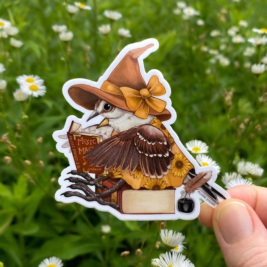 Sticker of a bird in a witch hat holding a book against a natural background with flowers.