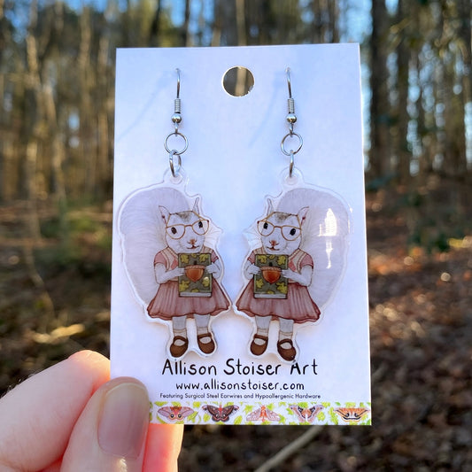 Earrings of white squirrels holding books on a card held by a hand, with a forest background.