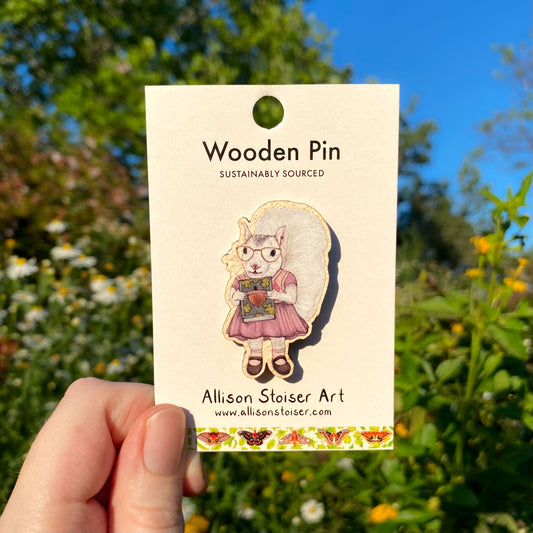 Wooden pin of a white squirrel holding a book on a card held in front of a natural background with greenery and flowers.