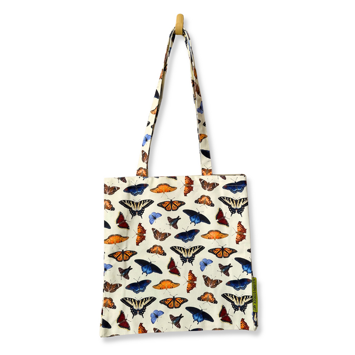 A canvas tote bag with a cream background and an all-over print of butterflies, hanging from a wooden peg on a white wall.