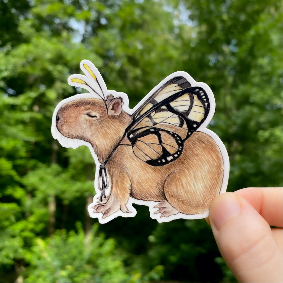 A hand holding a weatherproof vinyl sticker of a capybara with amber clearwing wings.