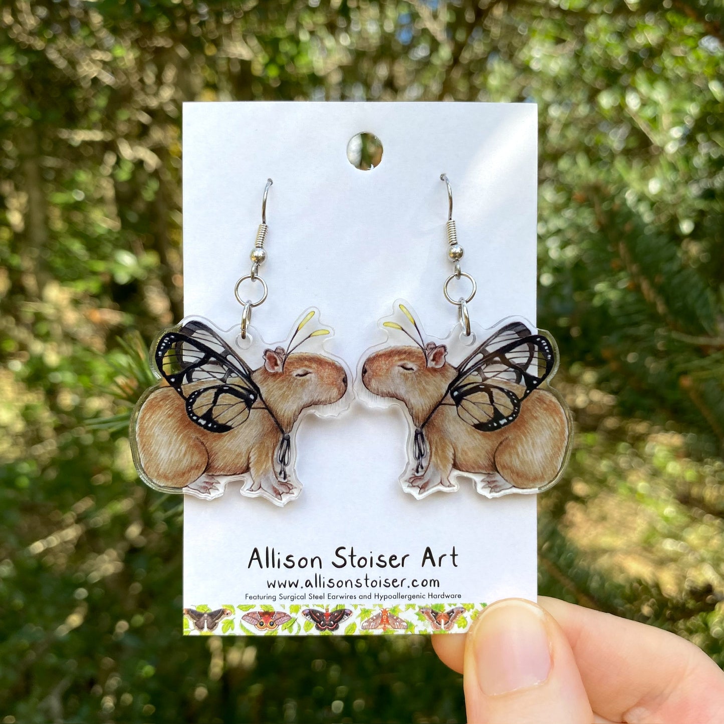 A hand holding a pair of butterfly capybara acrylic earrings