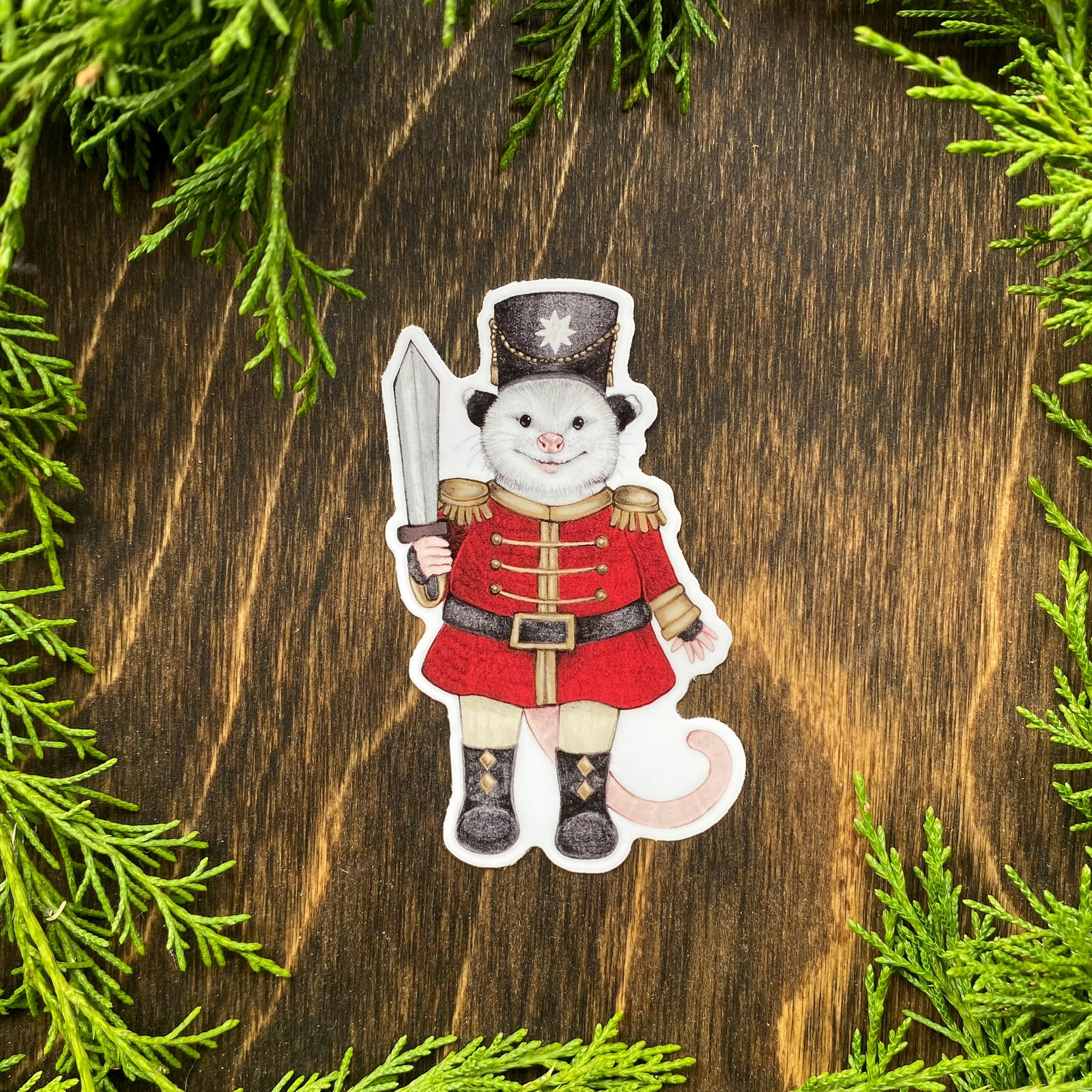 A weatherproof vinyl sticker of an opossum dressed in a red nutcracker outfit surrounded by cedar foliage.