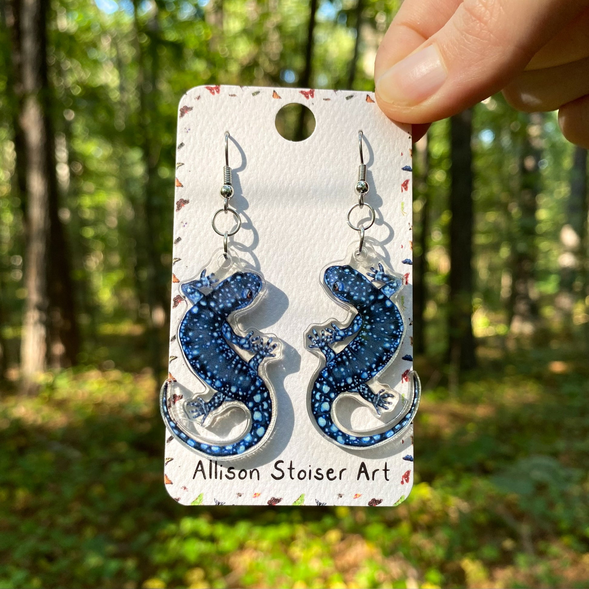 A hand holding a pair of acrylic blue-spotted salamander earrings.