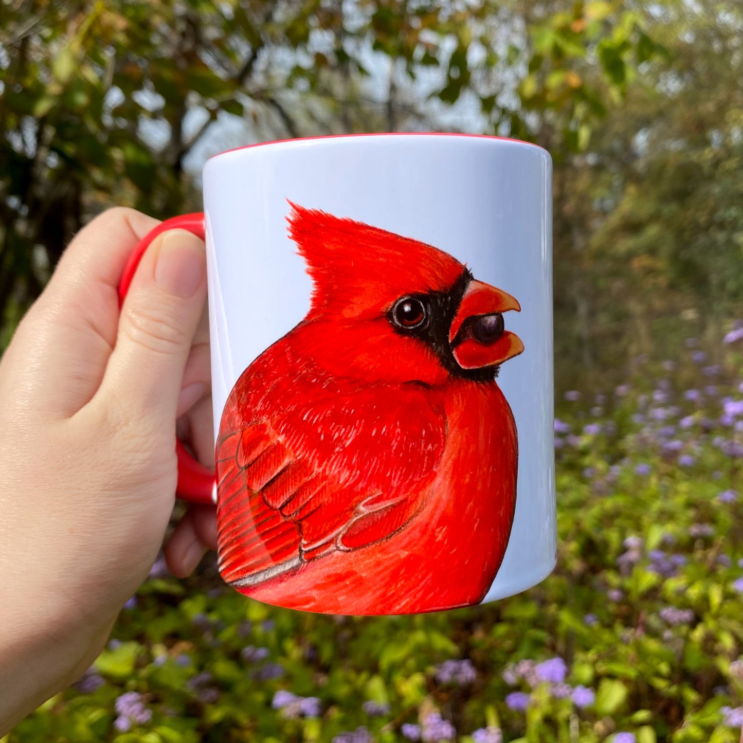 White mug with a male cardinal illustration held in front of a natural background with greenery and flowers.