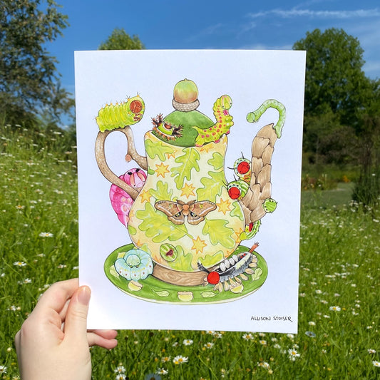 A hand holding a print of a green teapot decorated with leaves and stars surrounded by caterpillars.