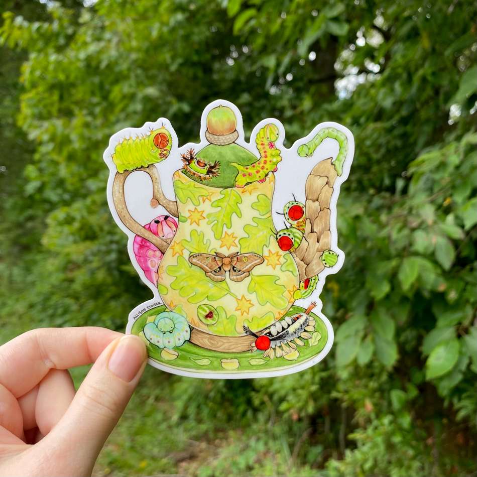 A hand holding a sticker of a green teapot surrounded by caterpillars.