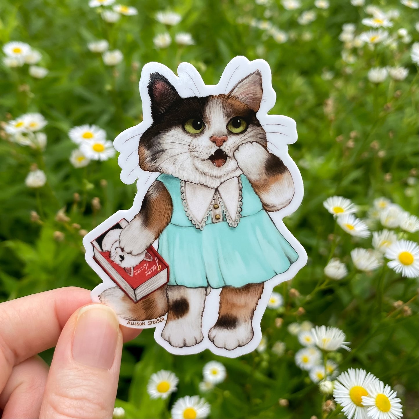 Sticker of a cat in a dress holding a book held in front of a field of daisies