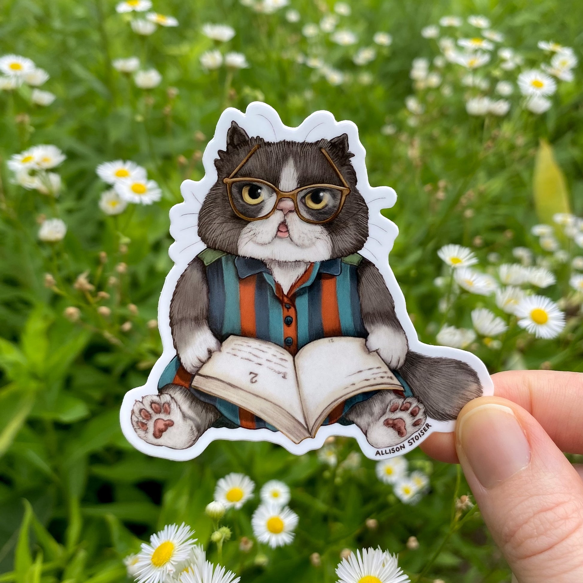 Sticker of a cat wearing glasses and reading a book held in front of a natural background with flowers.