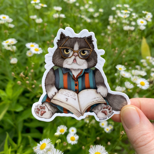 Sticker of a cat wearing glasses and reading a book held in front of a natural background with flowers.