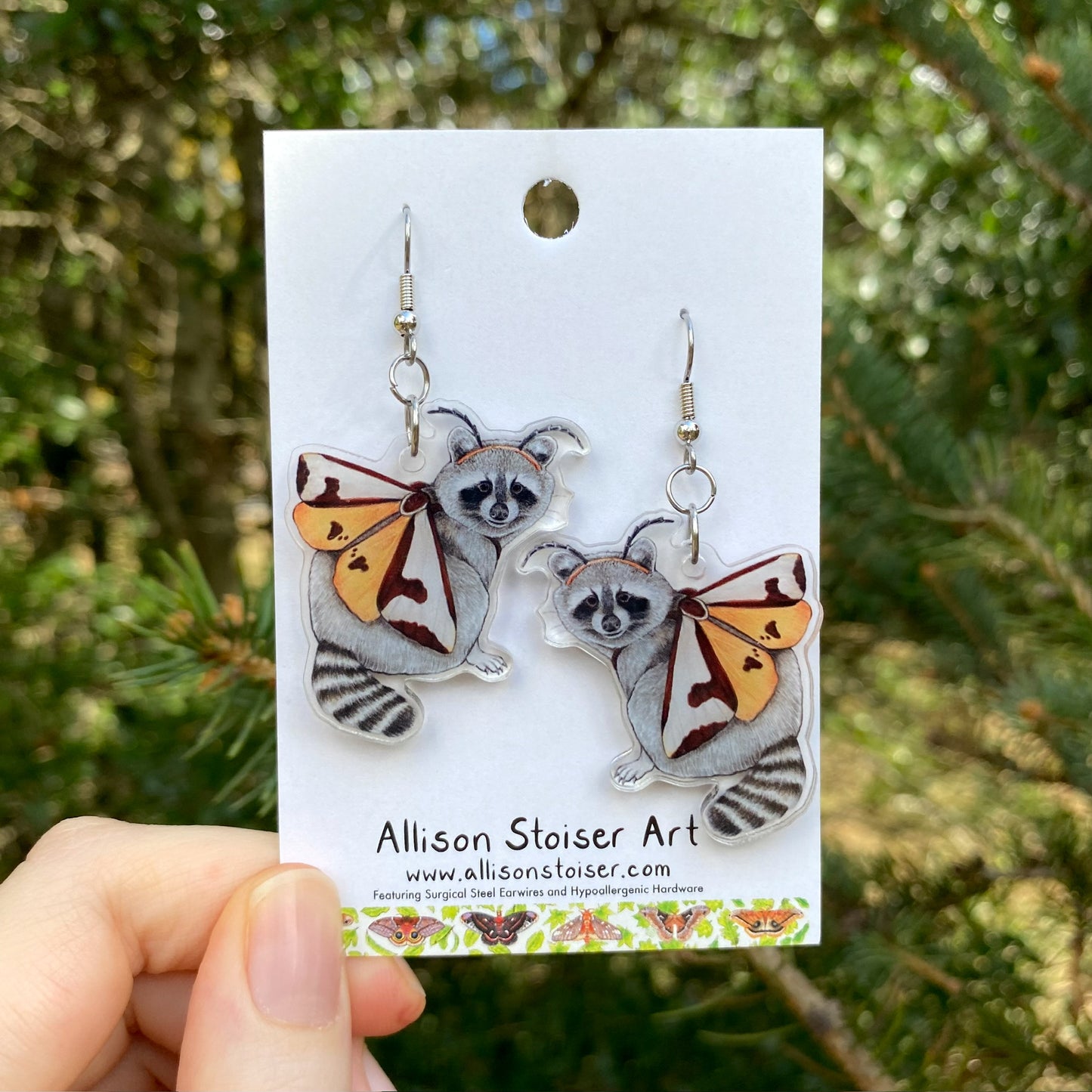 A hand holding a pair of earrings with charms of raccoons dressed up as moths.