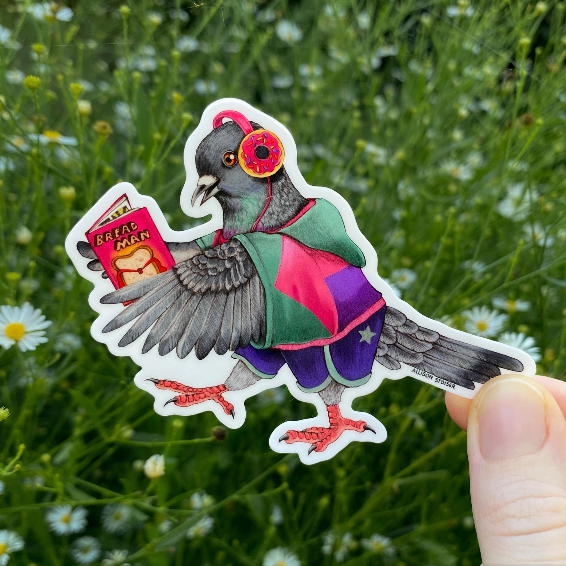 A die-cut sticker featuring an illustrated pigeon dressed in a teal and pink 90s inspired windbreaker holding a comic book, held by a hand against a blurred green field of daisies.