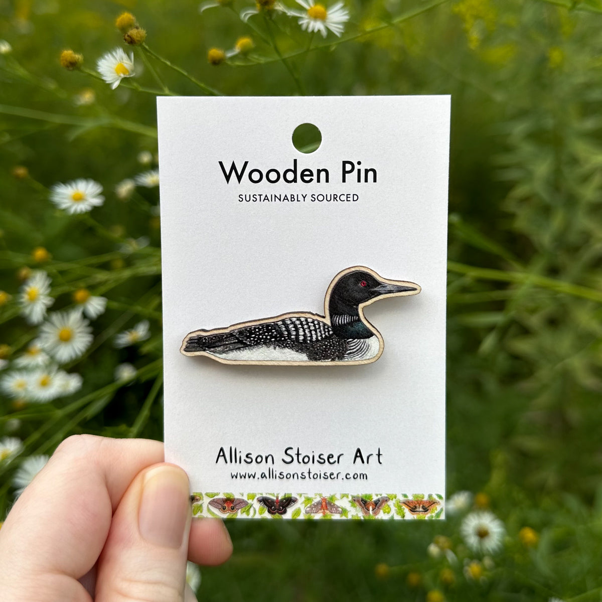 A wooden pin shaped like a loon, displayed on a white backing card held by a hand against a blurred background of green foliage and small white asters. The backing card reads 'Wooden Pin' and 'Allison Stoiser Art'.