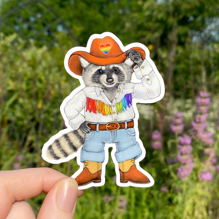 A hand holding a vinyl sticker of a raccoon dressed up in a rainbow cowboy outfit.