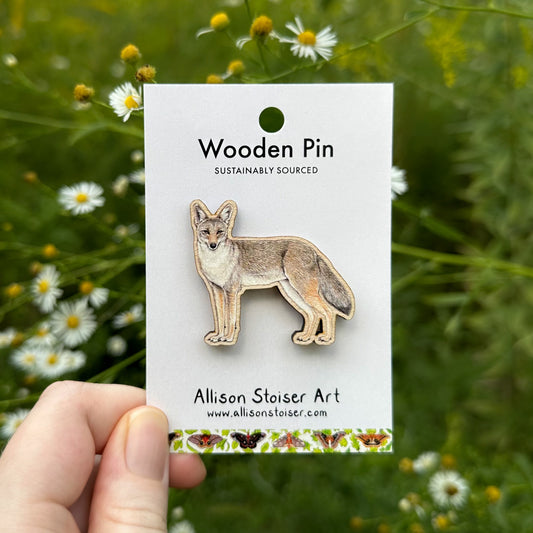 A hand holds a white display card featuring a wooden coyote pin. The card reads 'Wooden Pin' and 'Allison Stoiser Art' with the website allisonstoiser.