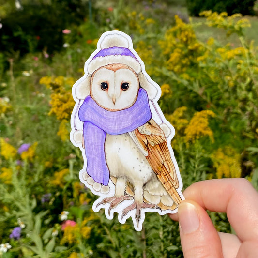 A vinyl sticker of a barn owl wearing a scarf and hat, held against a floral background.