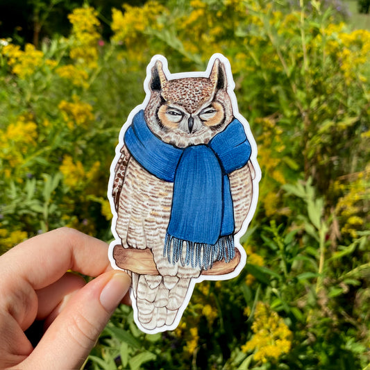 A vinyl sticker of a great-horned owl wearing a blue scarf, held against a background of yellow flowers.