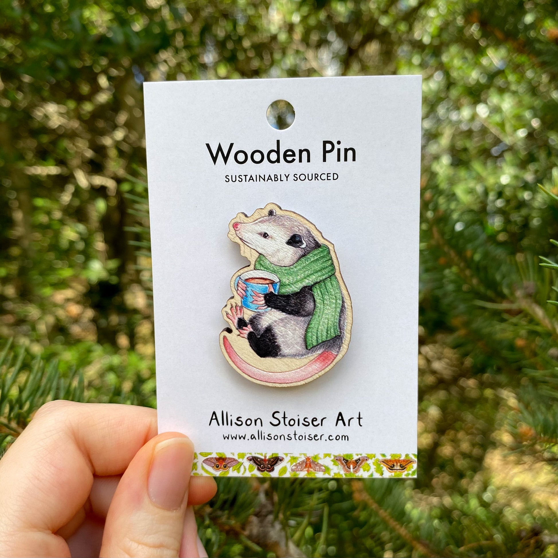 A hand holding a wooden pin of a cozy opossum dressed up in a green scarf