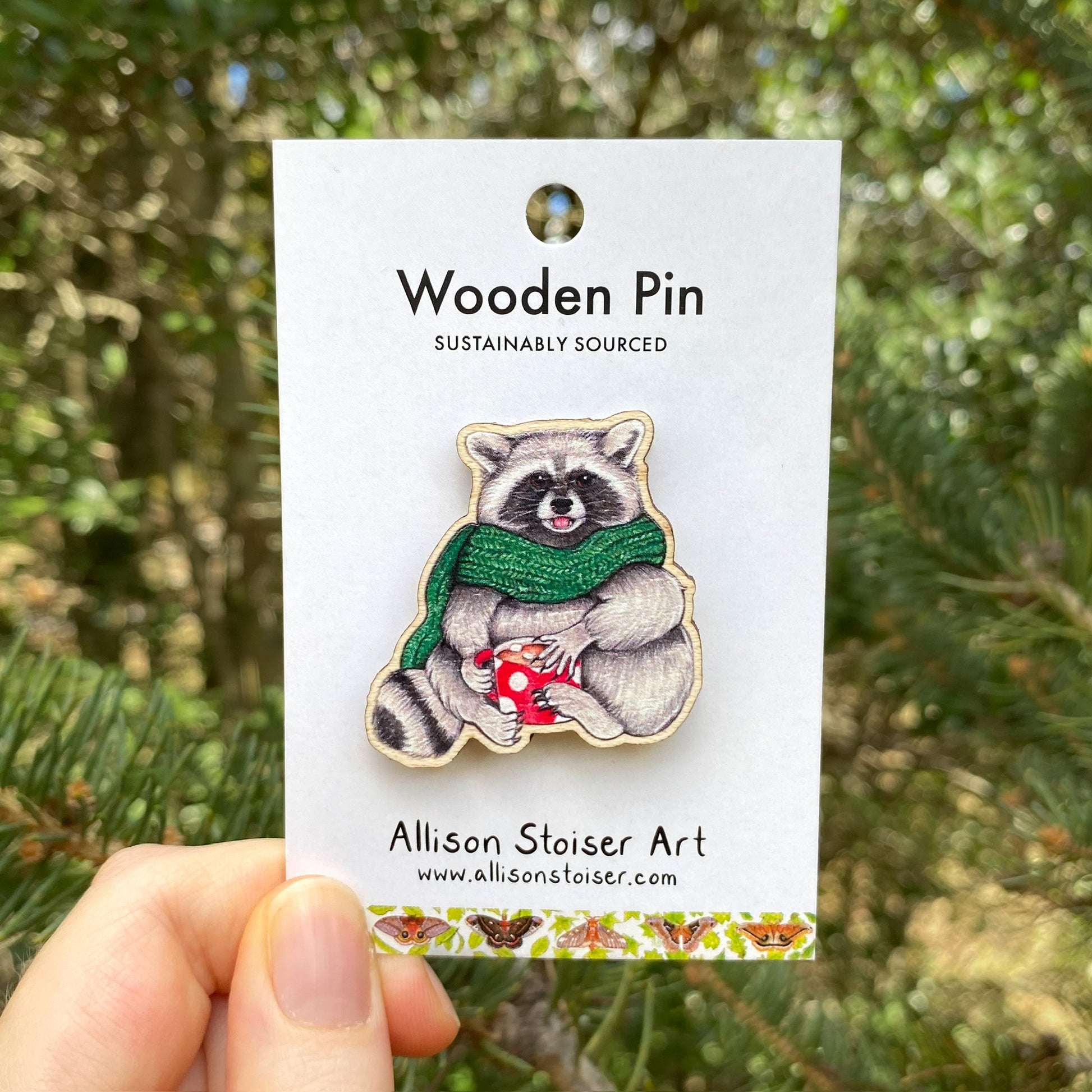 A hand holding a wooden pin of a raccoon dressed up in a green scarf with a mug of hot chocolate.