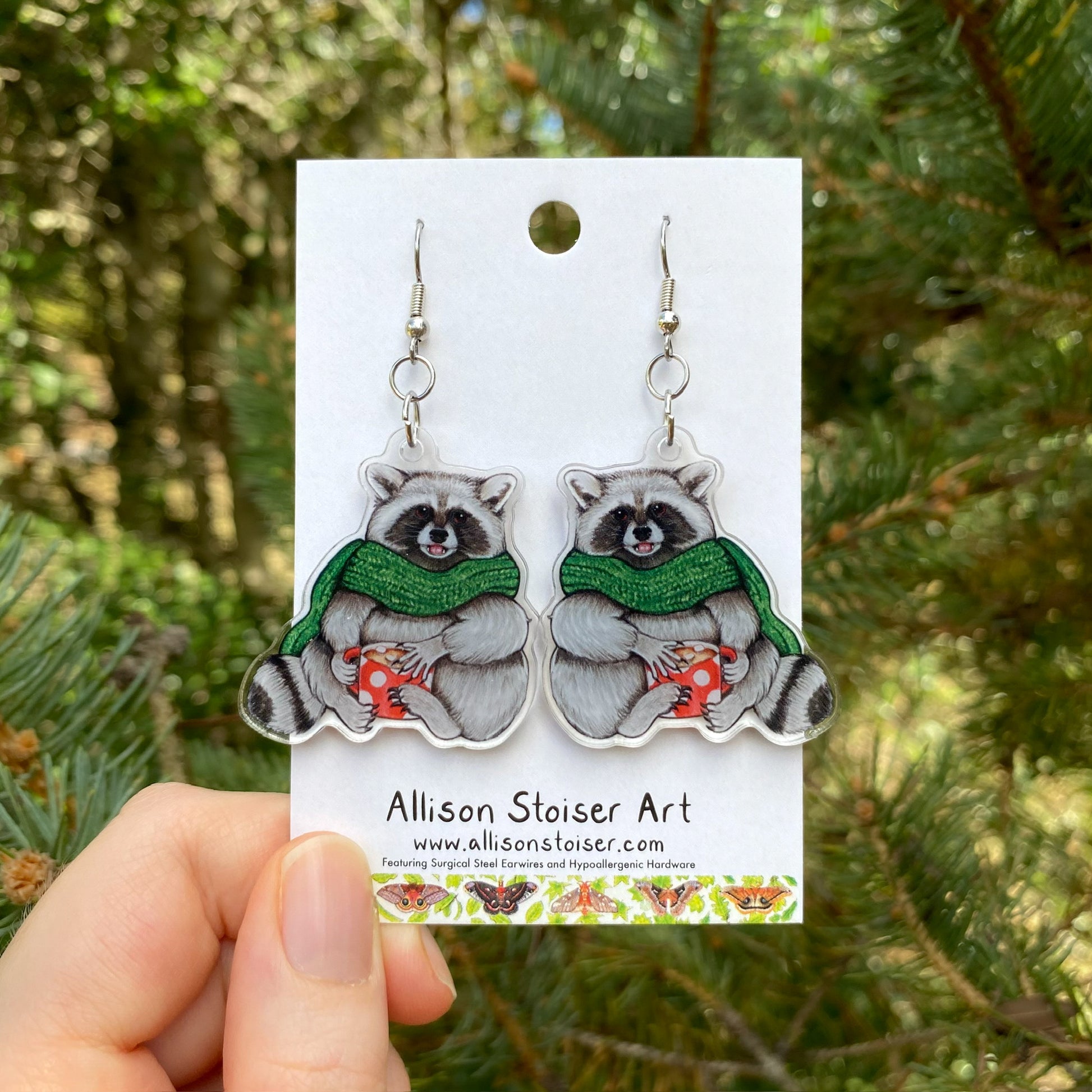 A hand holding a pair of cozy raccoon with scarf acrylic earrings