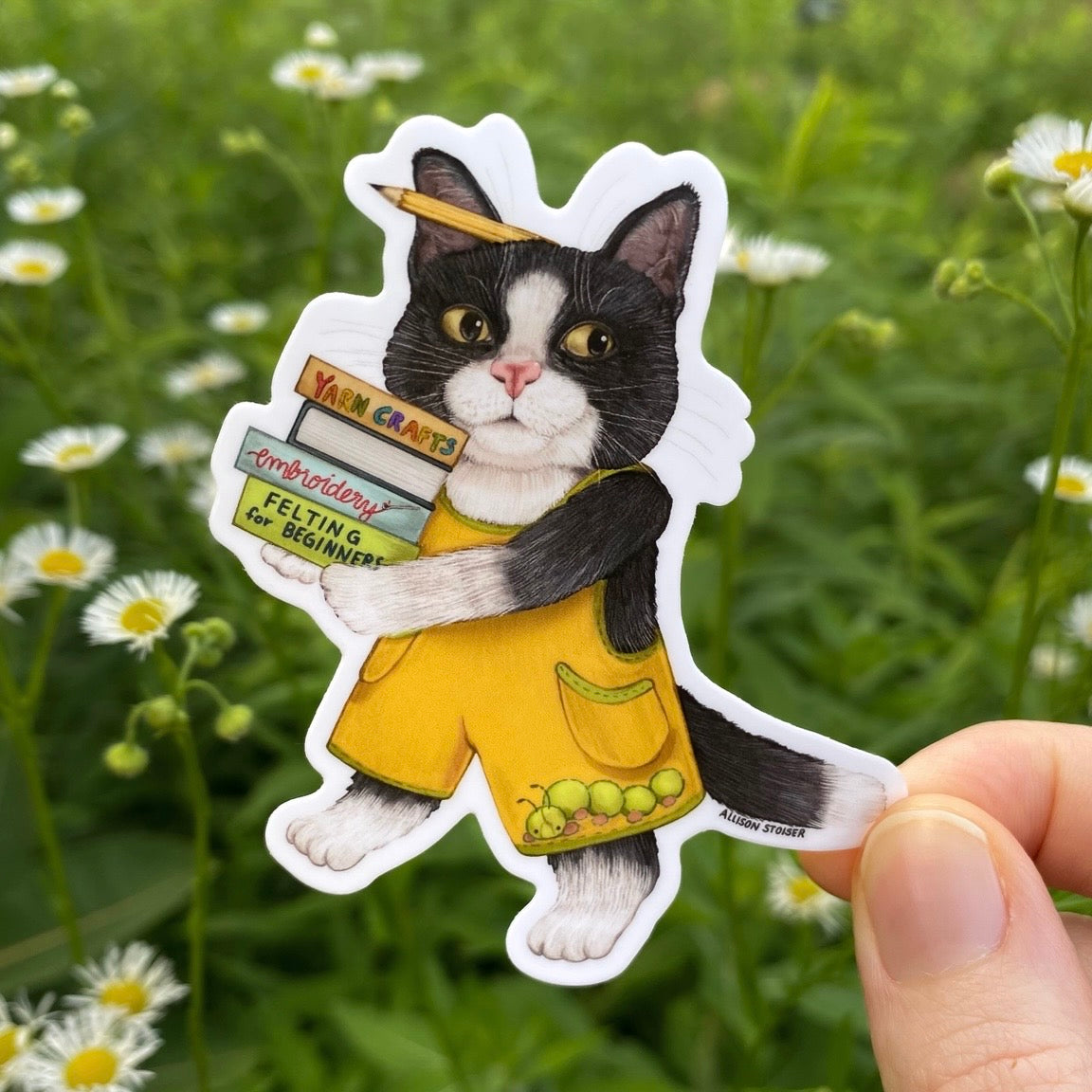 Sticker of a cat in yellow overalls holding books against a natural background