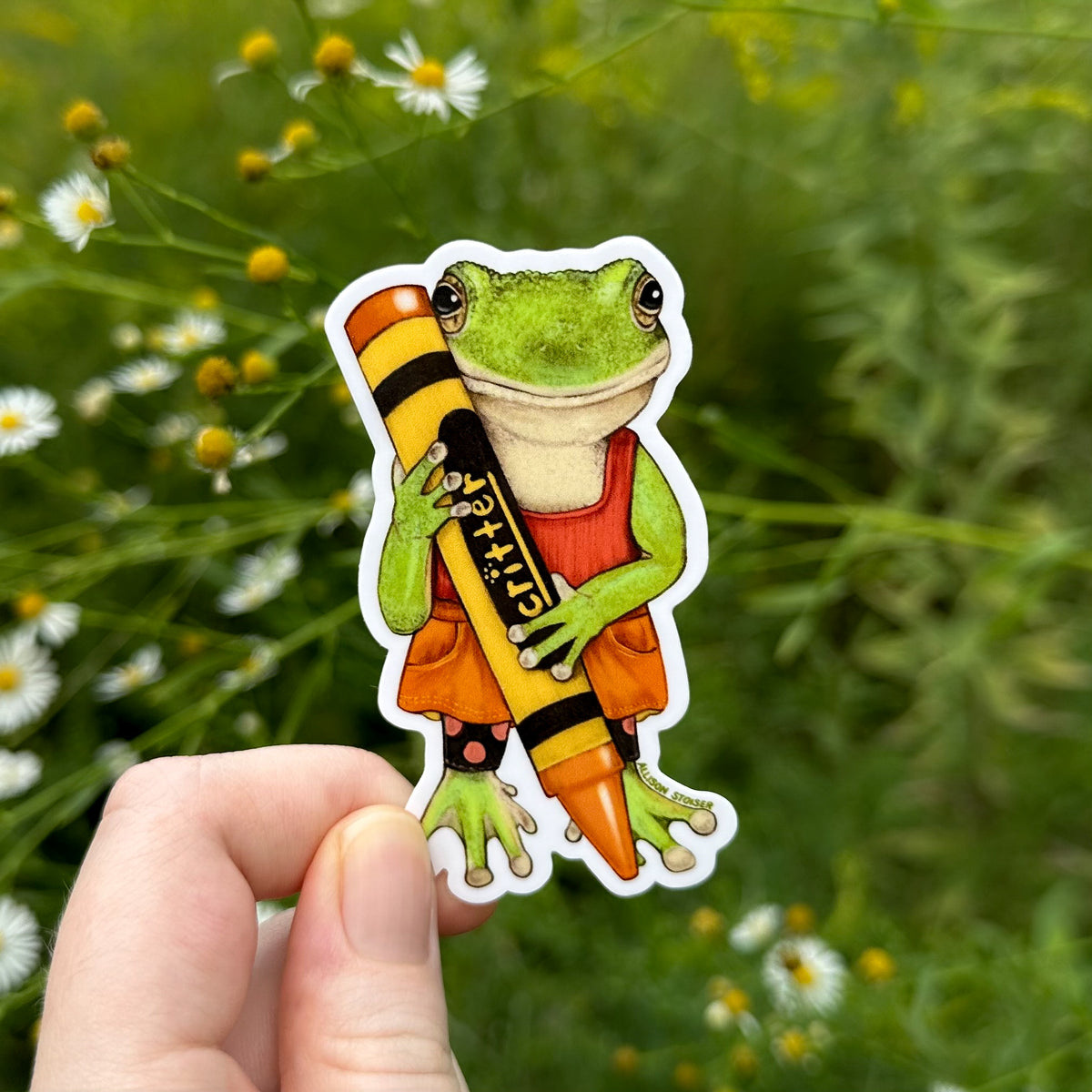 A die-cut sticker featuring a green tree frog holding an orange crayon, held by a hand against a blurred background of green plants and asters.