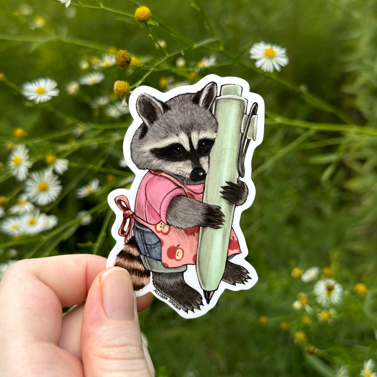 A hand holds a die-cut sticker featuring an illustrated raccoon in a pink shirt holding a large green pen, set against a blurred outdoor background of green foliage and asters.