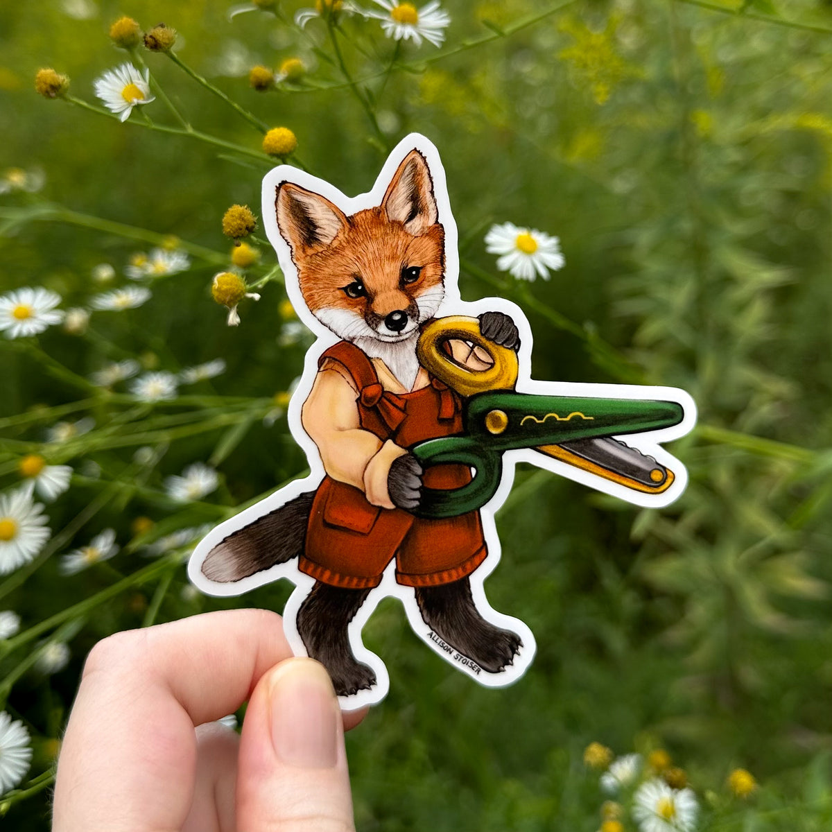 A die-cut sticker featuring an illustrated fox holding green and yellow craft scissors, held by a hand against a blurred outdoor background of green foliage and asters.