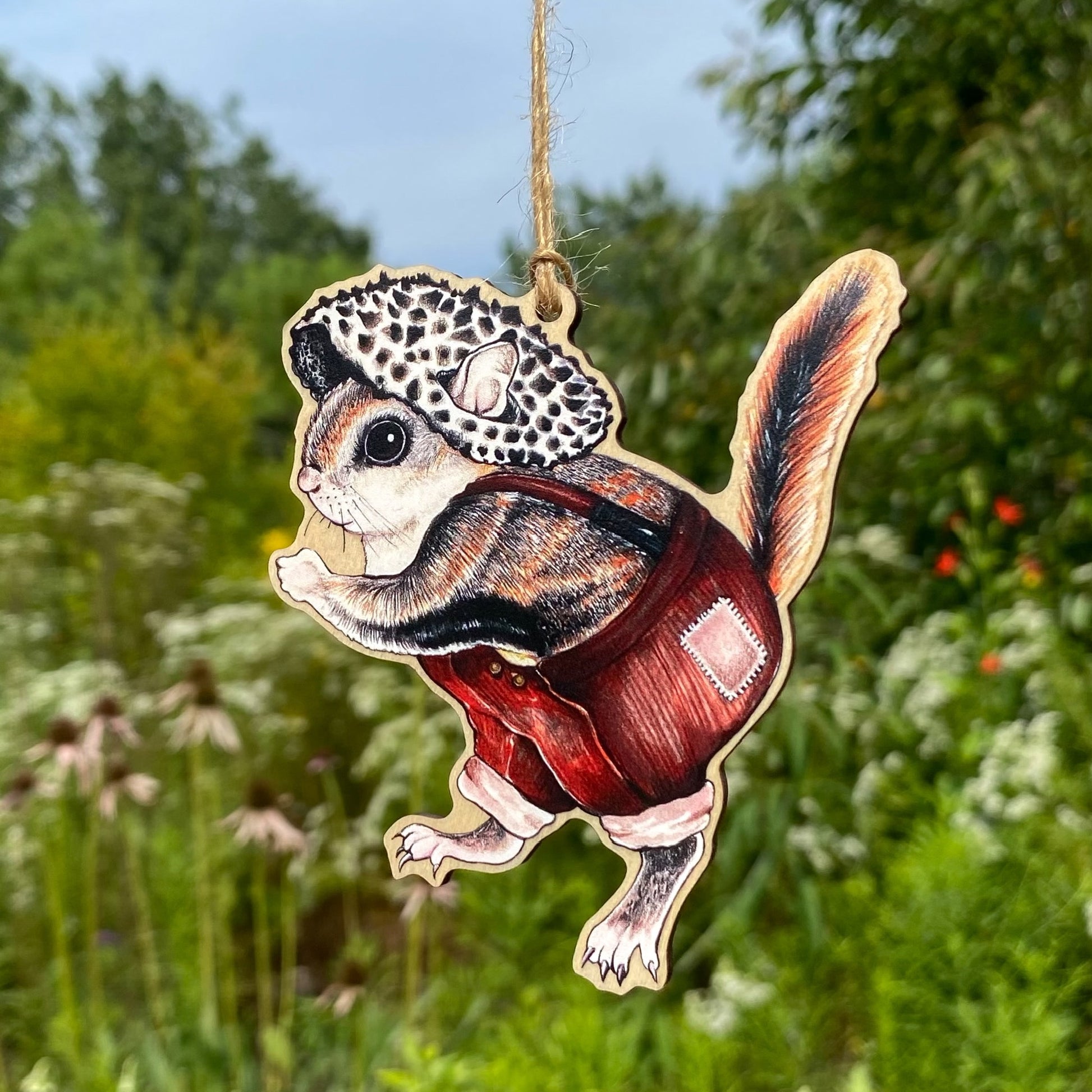 A wooden ornament strung with twine of a marching flying squirrel wearing overalls and an old man of the woods mushroom hat.