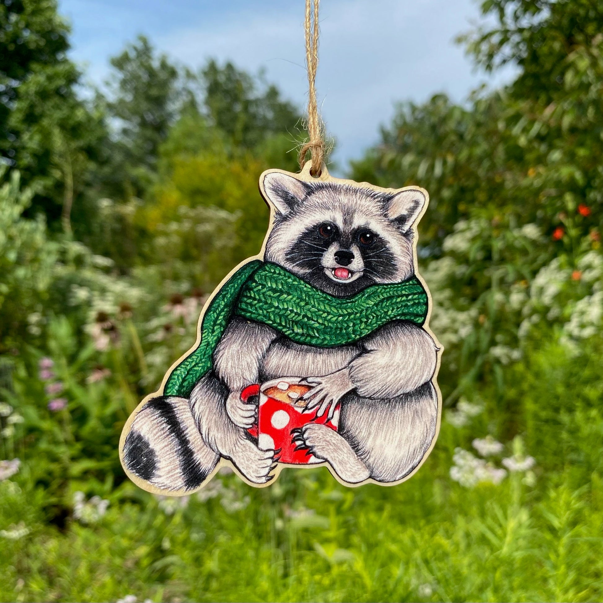 A wooden ornament strung with twine of a seated raccoon in a green scarf and holding a mug of hot chocolate.