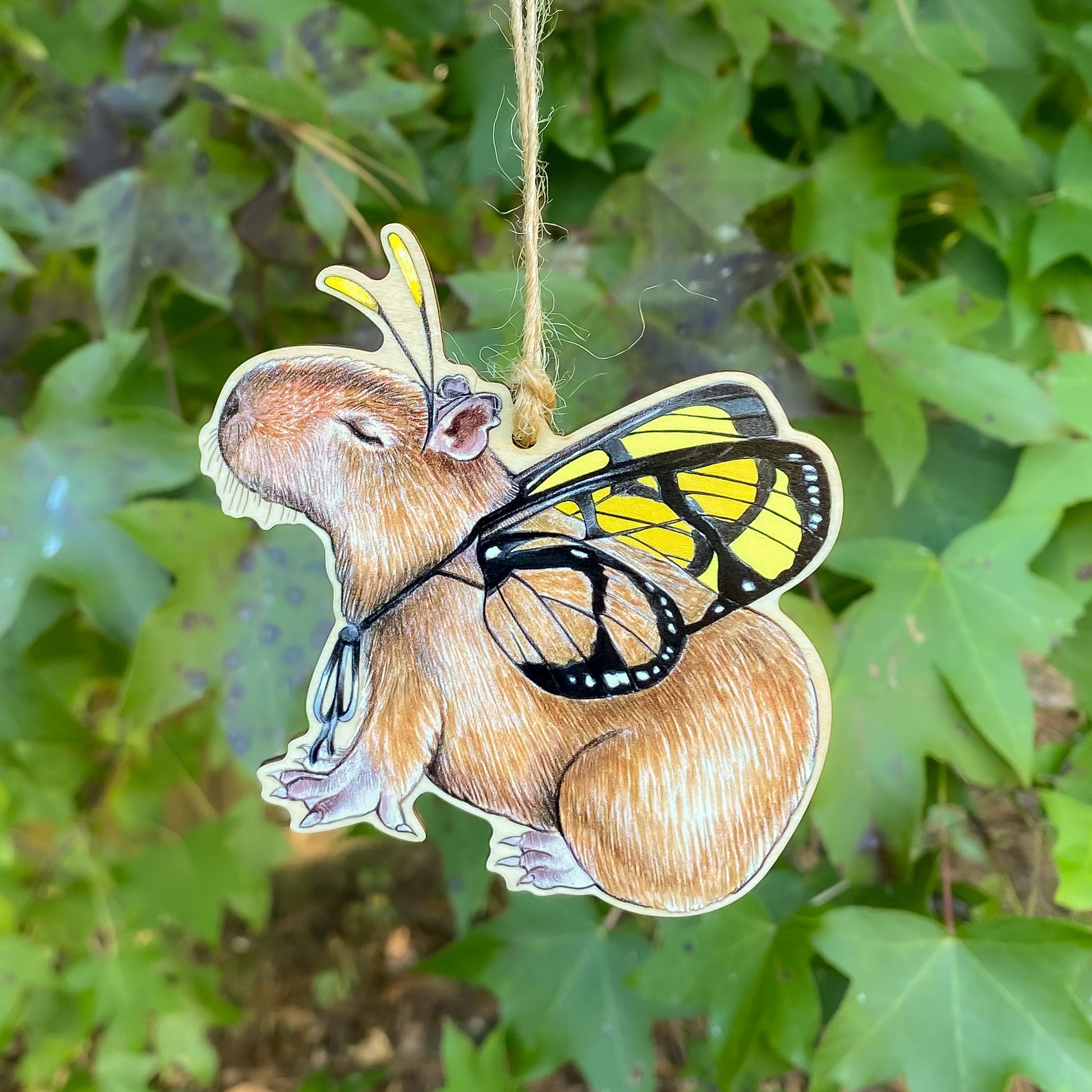 A wooden ornament strung with twine of a capybara with amber clearwing butterfly wings.