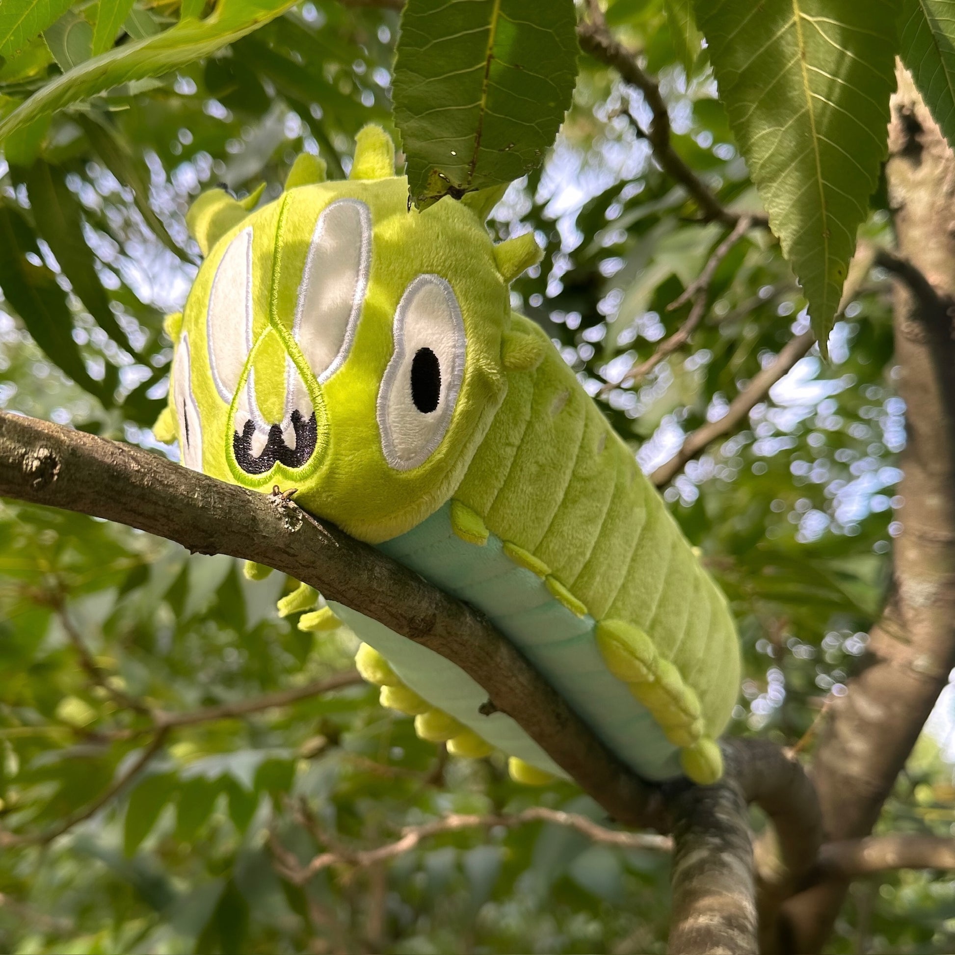 A green caterpillar plush toy with embroidered eyes and mouth, resting on a tree branch outdoors among green leaves.
