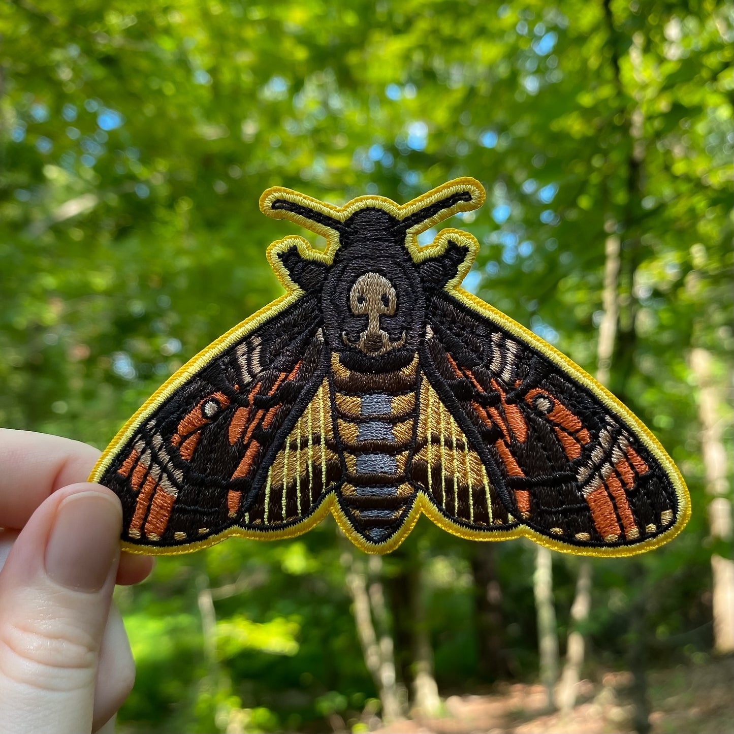 Embroidered death's head hawk moth patch held in front of a forest background