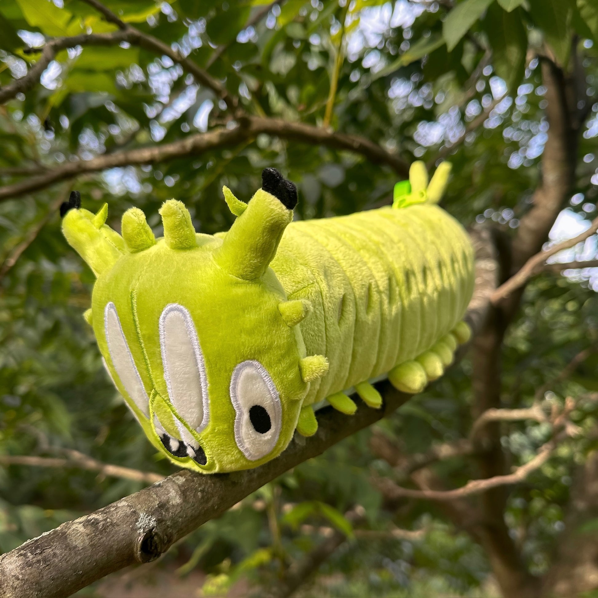 A bright lime green caterpillar plush toy with embroidered eyes and black-tipped antennae, resting on a tree branch outdoors with green leaves and branches blurred in the background.