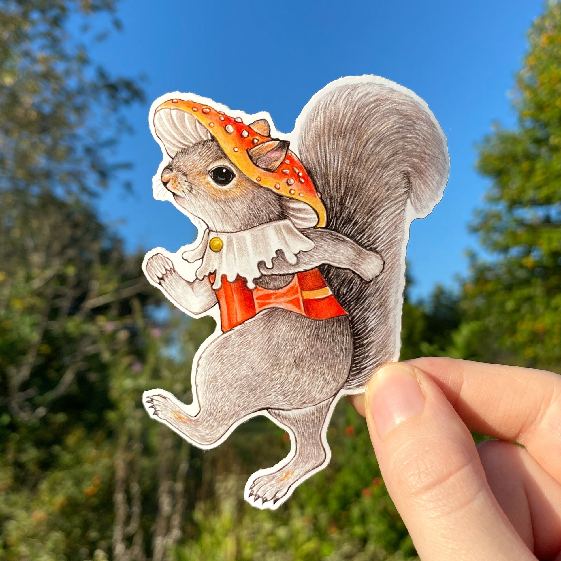 A hand holding a weatherproof vinyl sticker of a marching gray squirrel with a mushroom hat.