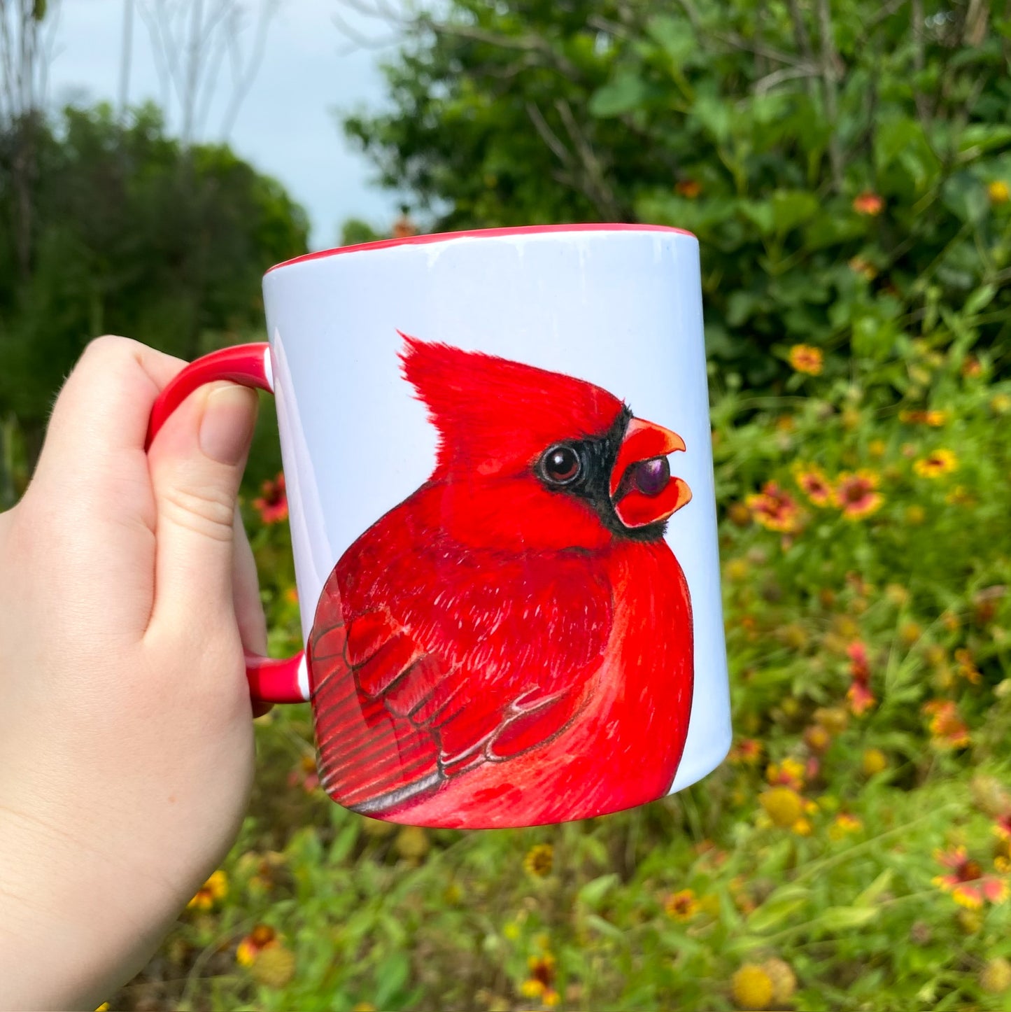 A hand holding a red-handled mug showing two North American cardinals. This image shows the side with the bright red male.