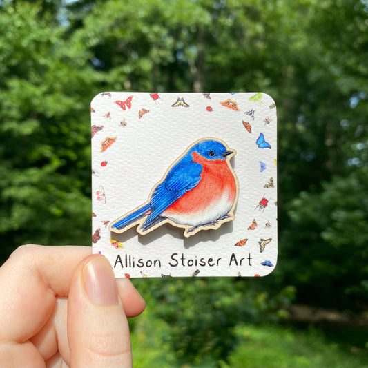 A hand holds a sustainably sourced wooden pin featuring an illustration of an eastern bluebird