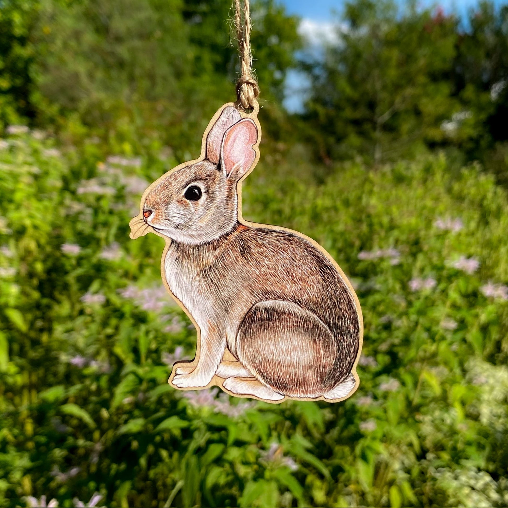 Wooden rabbit ornament hanging against a natural background with greenery and flowers.